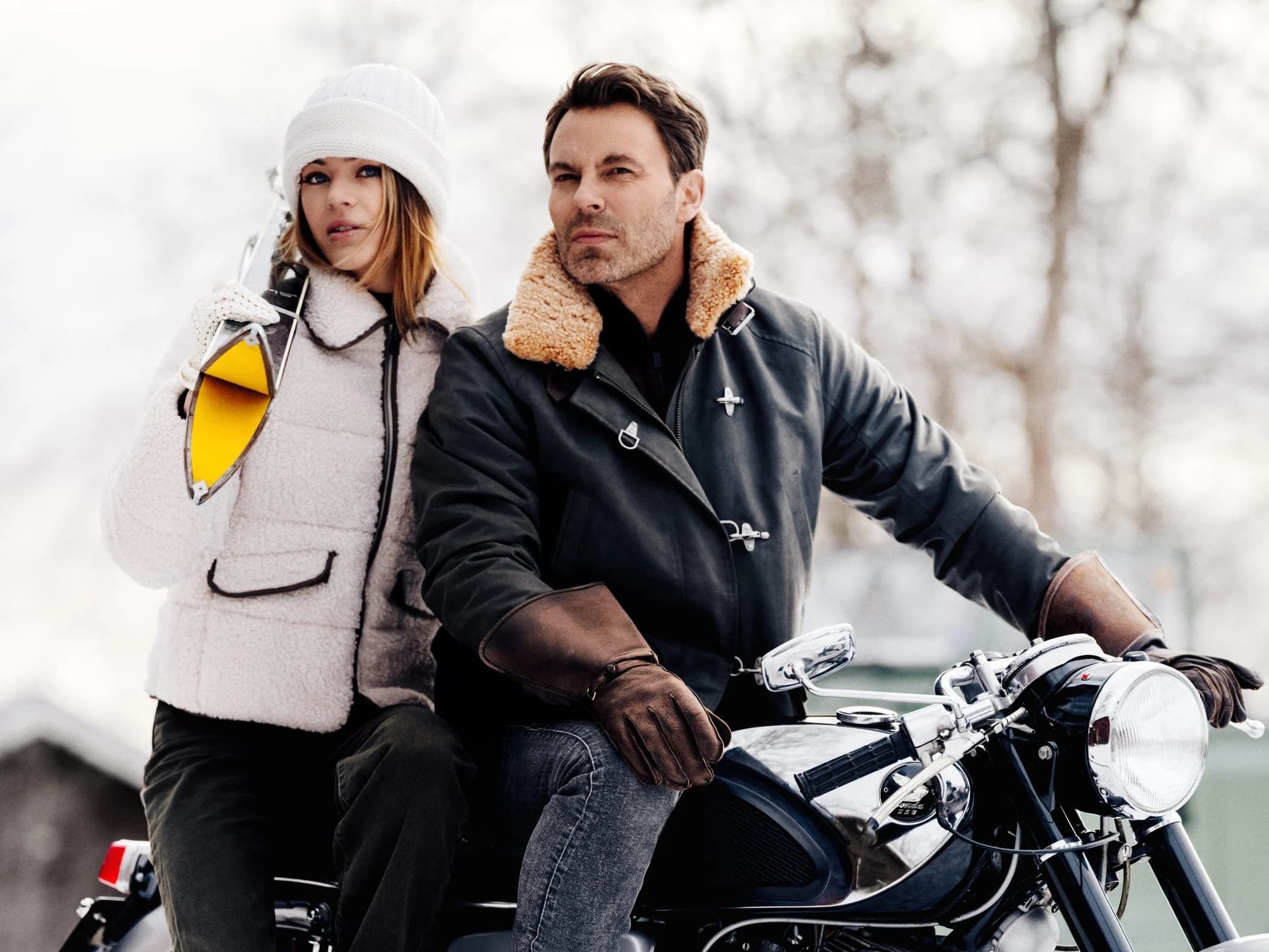 Two figures in winter outerwear beside a classic motorcycle in bare woodland