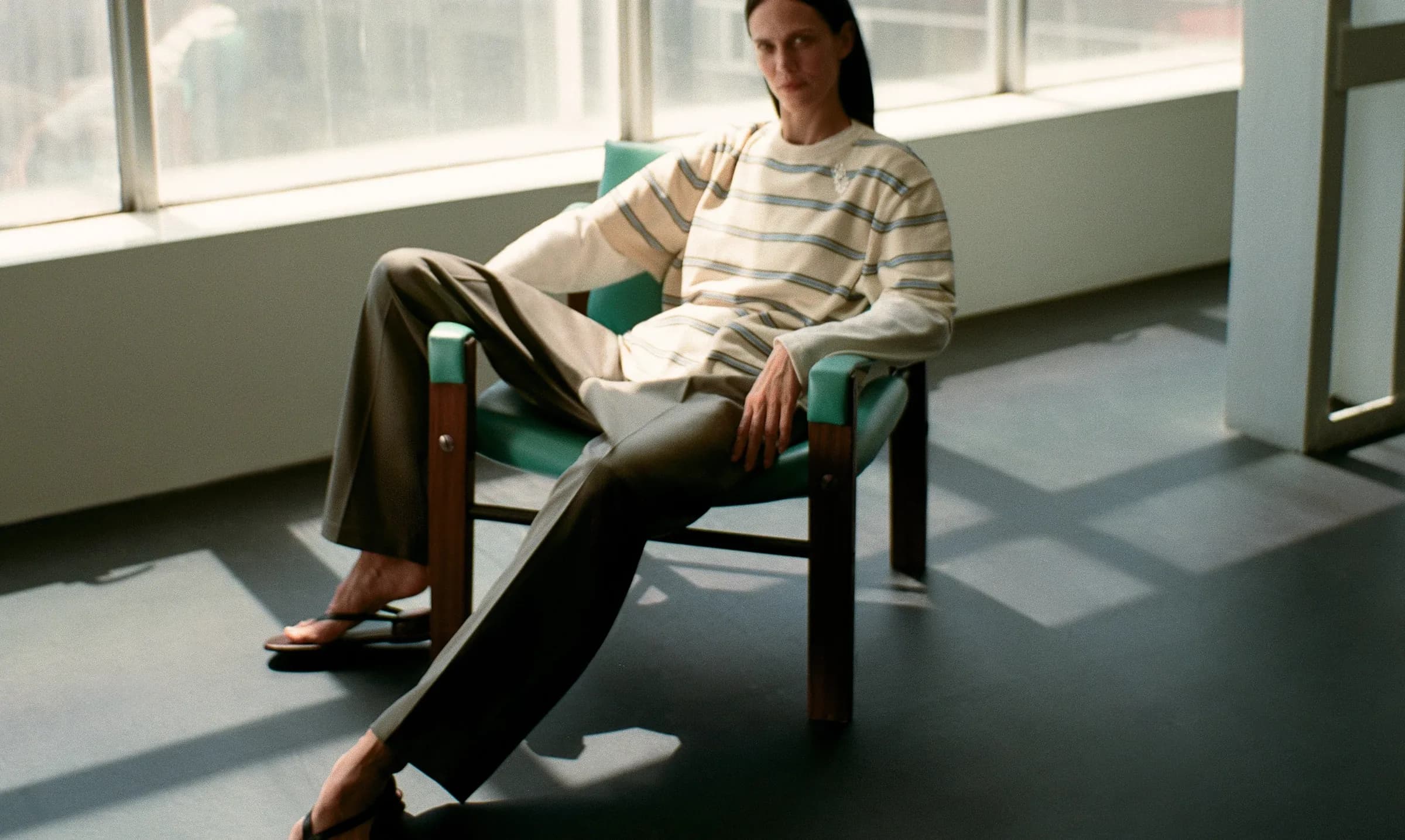A seated figure in soft tailoring beside a sunlit window wall