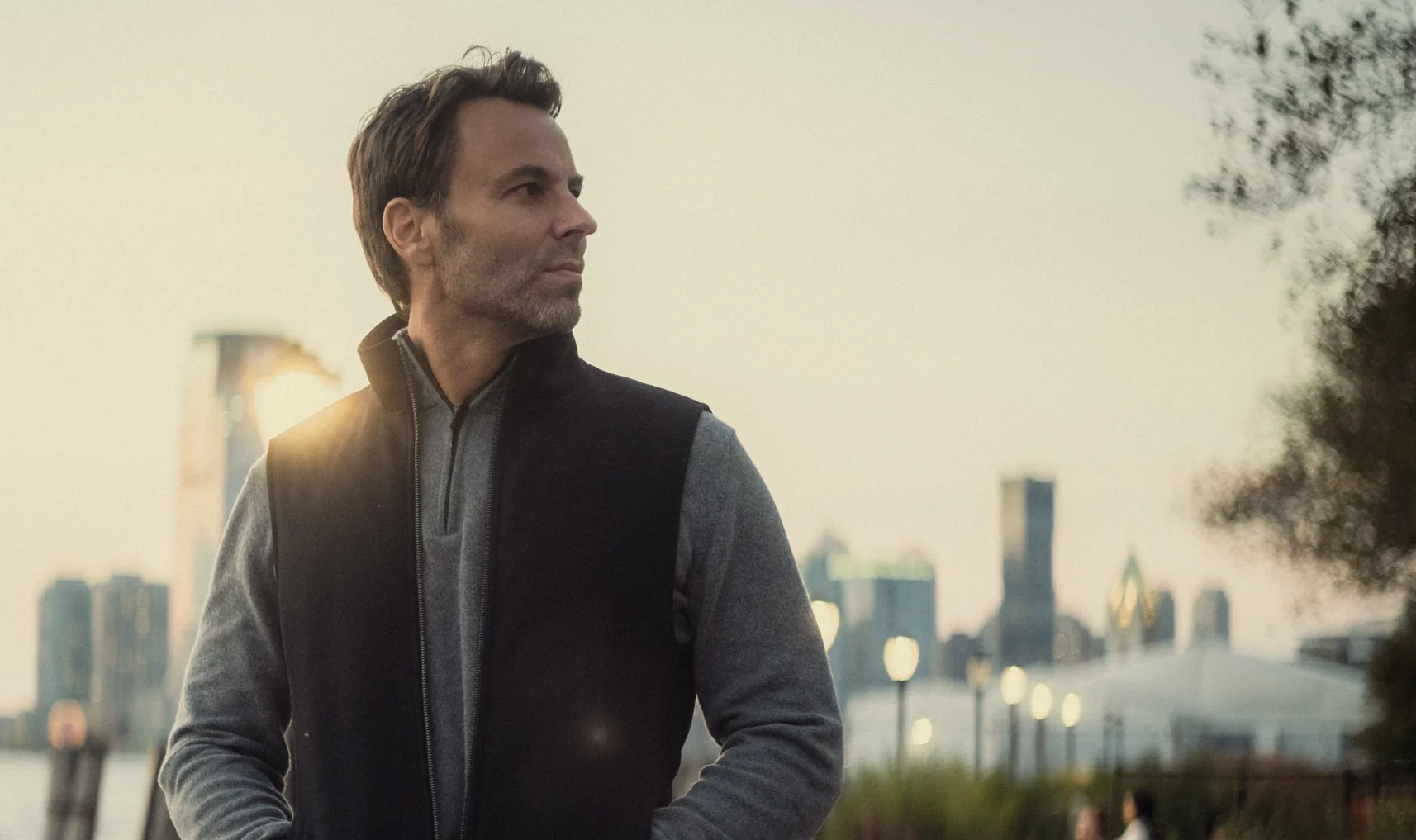 A figure in a technical gilet standing against a wide waterside skyline at dusk