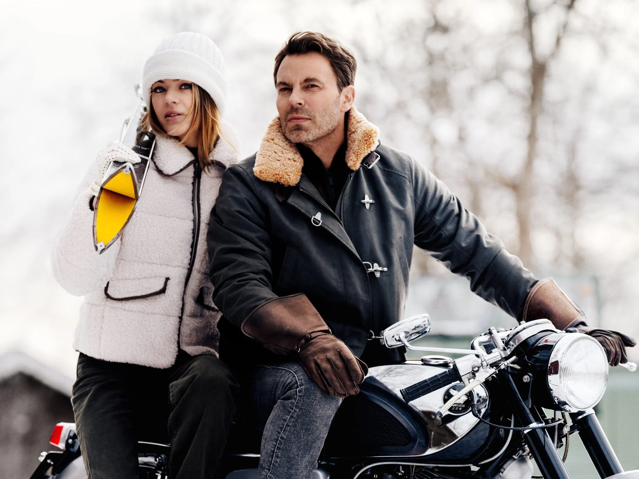 Two figures in winter outerwear beside a classic motorcycle in bare woodland