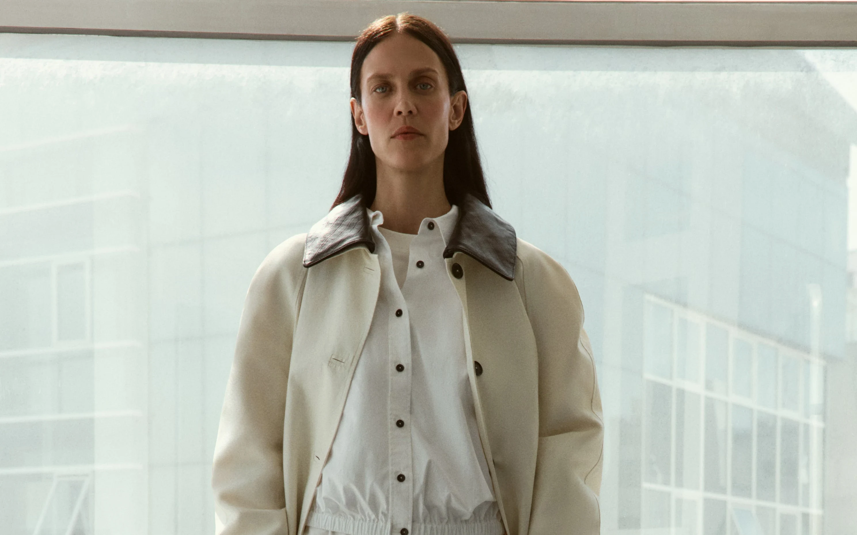 A figure in a pale coat and layered shirting against a bright glazed facade