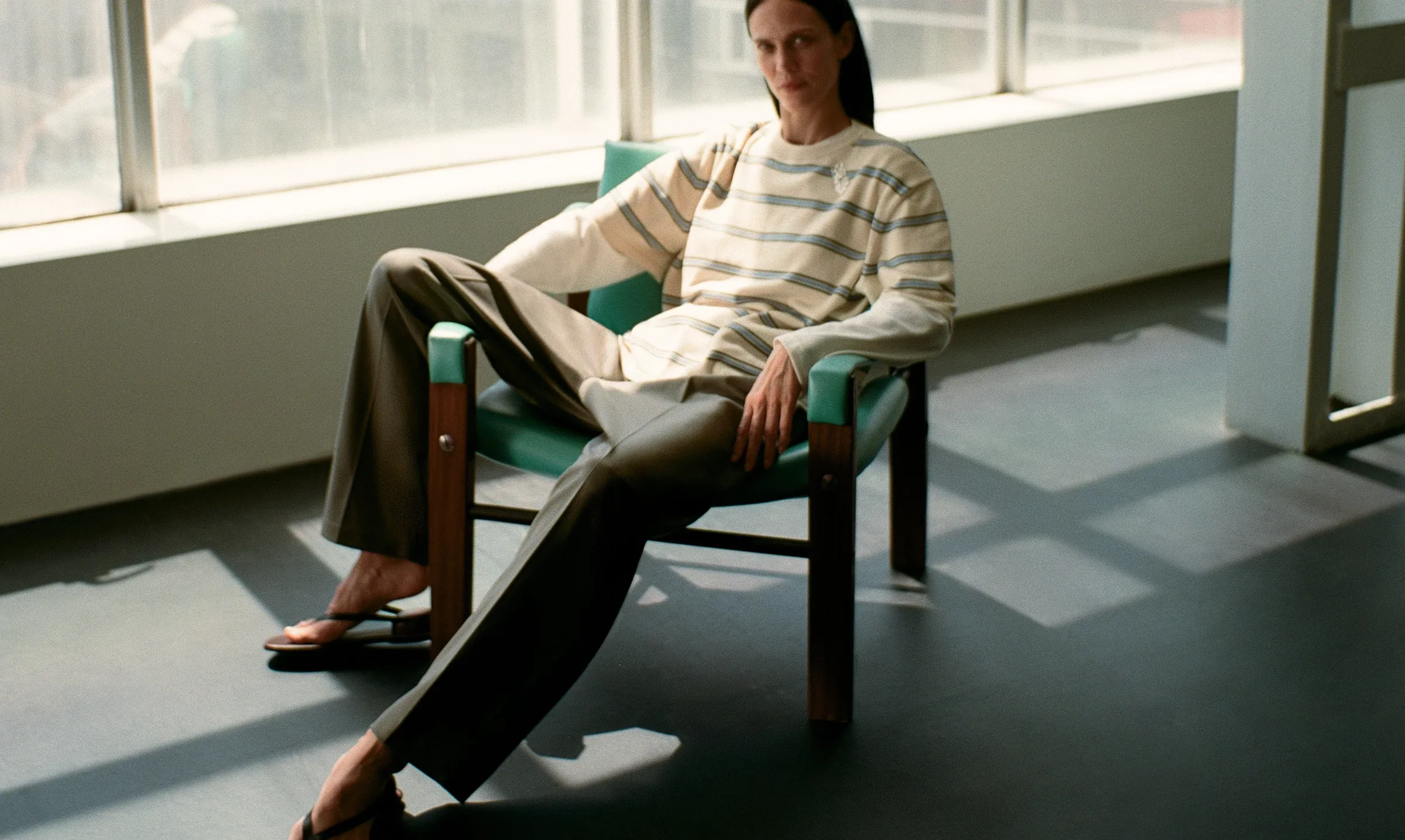 A seated figure in soft tailoring beside a sunlit window wall