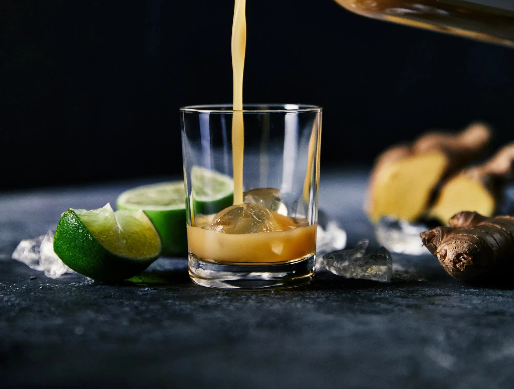 A dark-toned pour into a cut glass with limes and ginger on a slate surface